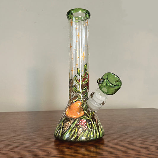 This is the front of the bong. The top rim and bowl are a matching forest green stained glass. There is a field of grass at the bottom with an orange mushroom and a small red with white dots mushroom. There are gold sparkles and dots going up the top. This is the front left view.