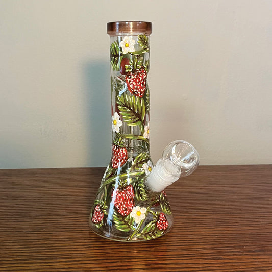 This is a beaker bong that is hand painted with a vine wrapping around the piece. There are strawberries, leaves, and little white flowers hanging off the vine. The top rim is a brownish red stained glass. This is the front view of the bong
