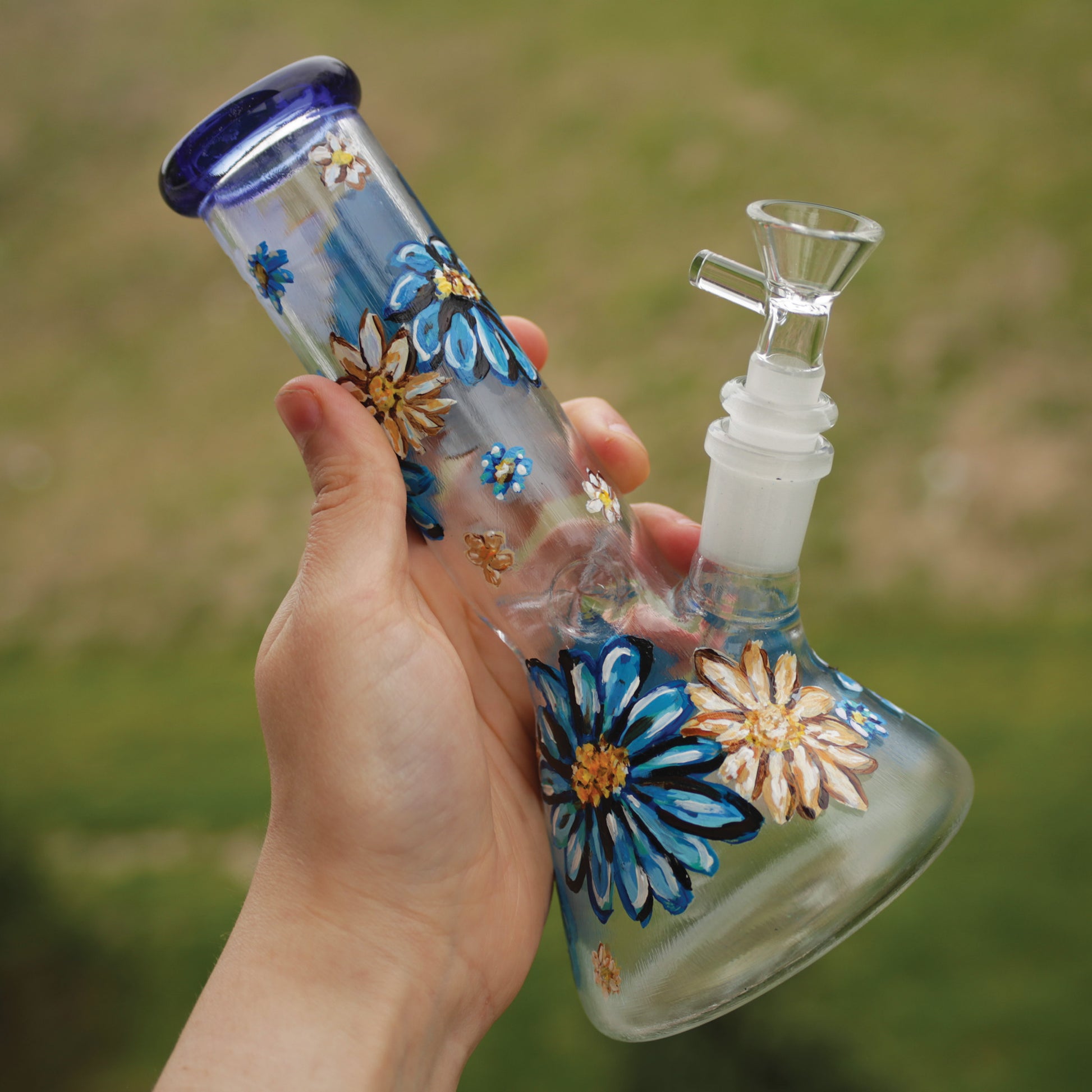 An outdoor picture of a 9 inch clear beaker bong with painted blue and white flowers with a blue rim top and ice catcher.